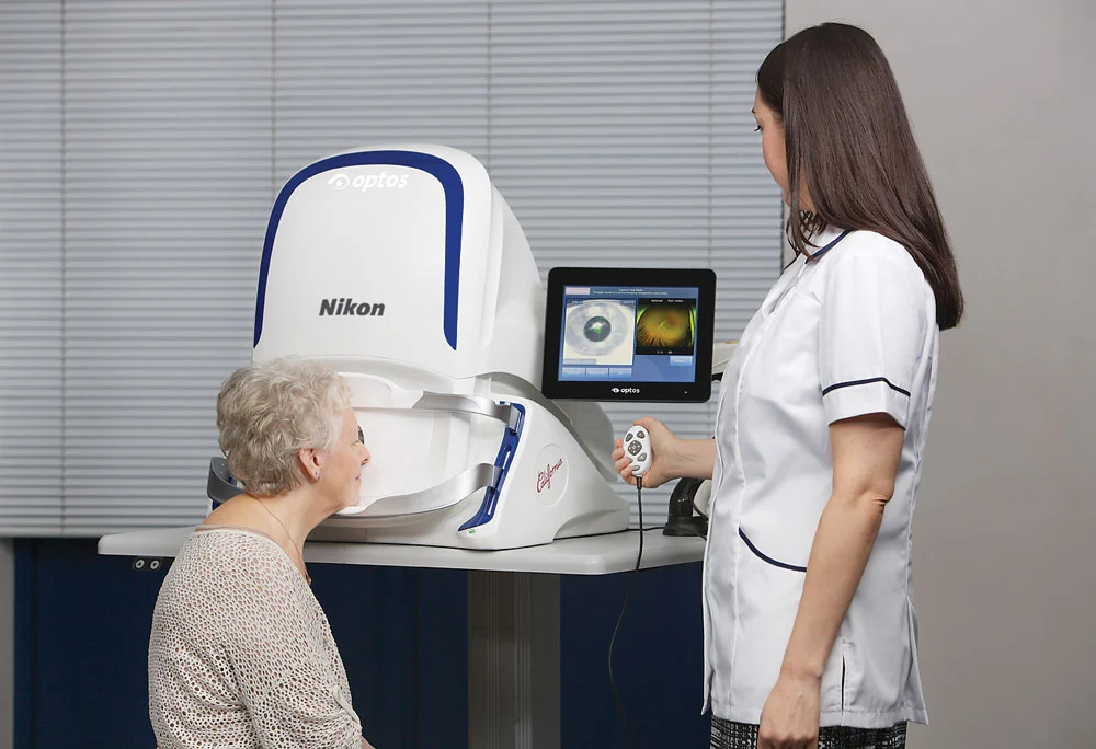 An optometry technician operates the Optos California ultra-widefield retinal imaging device while an older adult patient rests at the headrest during a scan